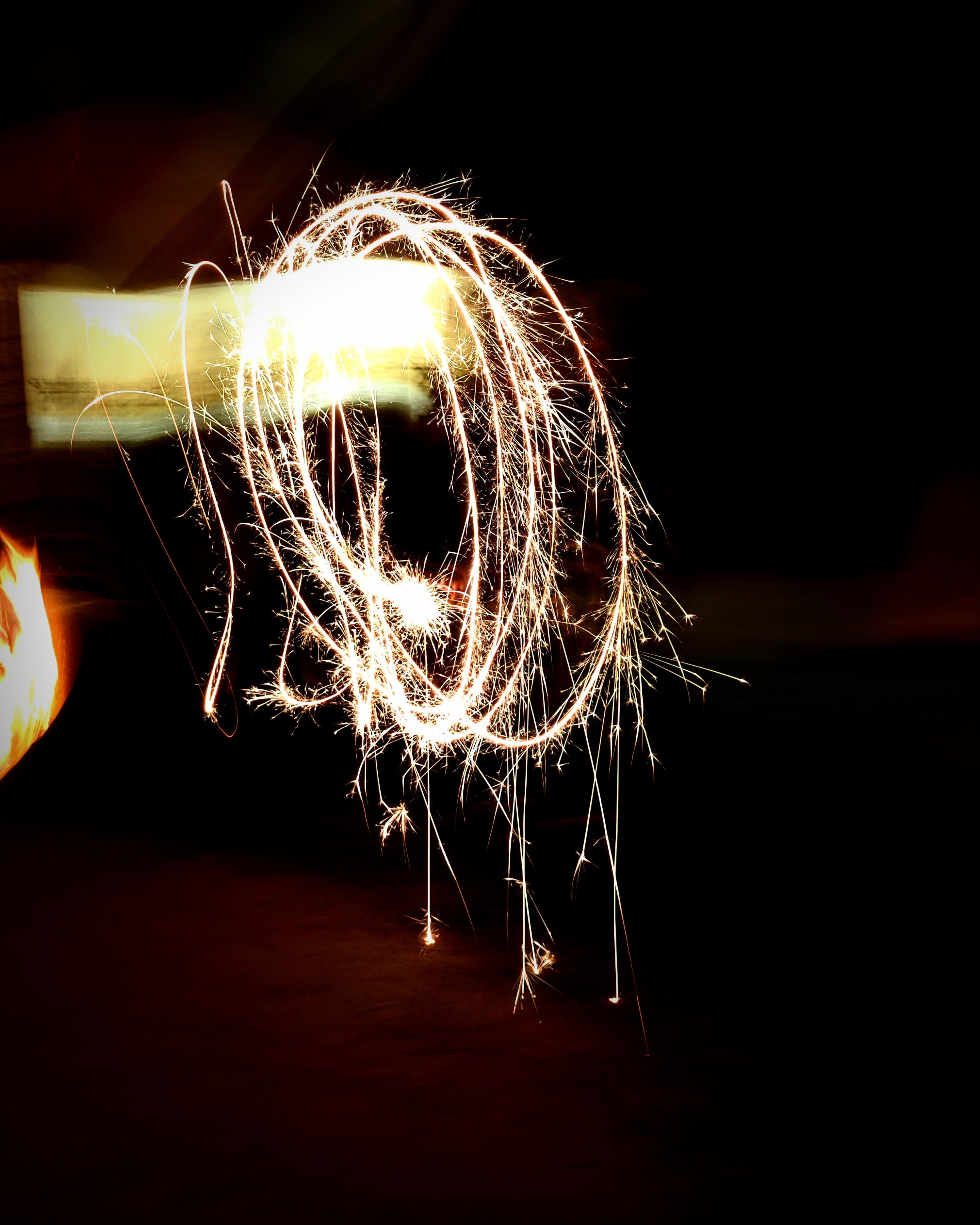 long-exposure shot of a sparkler being spun ain a circle, some bright light in the top left orner causing a streak that travels to the middle of the image